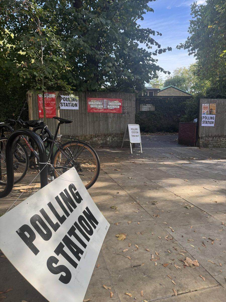 A 'Polling Station' sign propped against a railing beside parked bicycles, outside a gated polling station in Holborn and St Pancras.